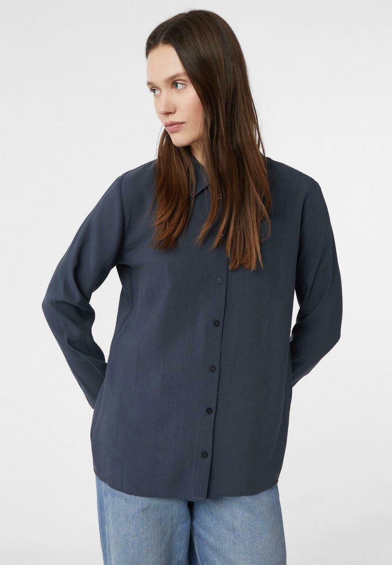 Granatowa koszula z długim rękawem, zapinana na guziki, z delikatną poziomą strukturą i klasycznym kołnierzykiem. Połączona z jasnoniebieskimi spodniami dżinsowymi.