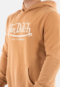 Sudadera color beige hecha de algodón con un bolsillo frontal. Cuenta con capucha ajustable con cordón y un logo impreso en blanco que dice "Von Dutch - CALIFORNIA." Textura suave.