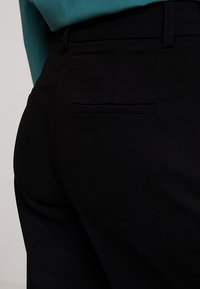 Fekete szövetnadrág sima felülettel, hátsó zsebbel és övhurkokkal. Az derékrész állítható a megfelelő illeszkedés érdekében.