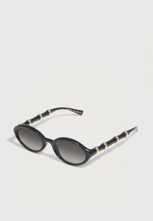 Lunettes de soleil ovales noires avec verres dégradés et branches de style bambou accentuées d'or, reposant sur une surface blanche.