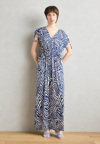 Robe maxi avec un motif zèbre abstrait bleu et crème, décolleté en V, taille élastique et manches courtes avec détails à nouer. Associée à des sandales bleu clair.