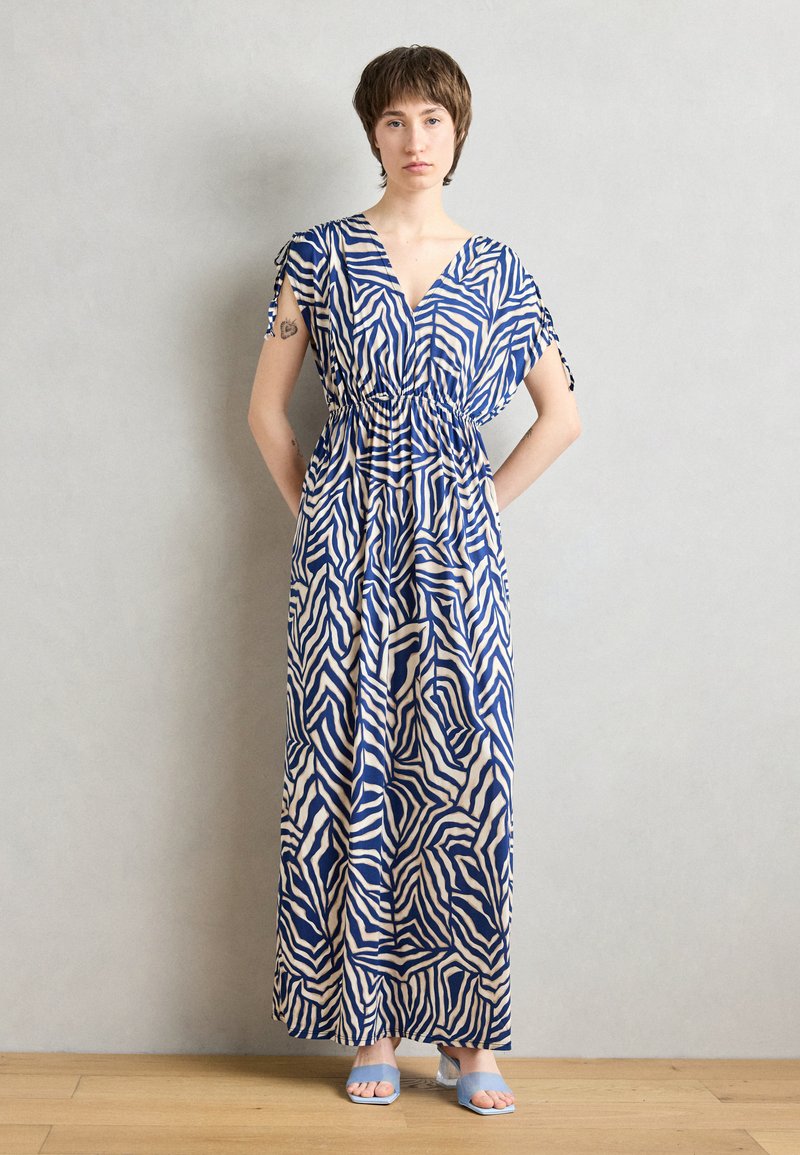 Robe maxi avec un motif zèbre abstrait bleu et crème, décolleté en V, taille élastique et manches courtes avec détails à nouer. Associée à des sandales bleu clair.