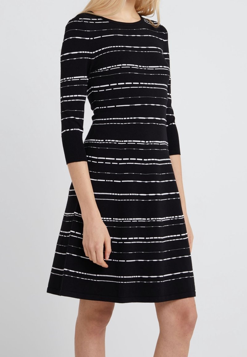 Femme portant une robe noire longueur genoux avec des manches trois-quarts et des motifs de rayures blanches horizontales discontinues.