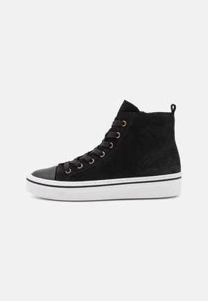 Czarny sneaker za kostkę z białą gumową podeszwą, czarnym czubkiem i czarnymi sznurowadłami na jednolitym jasnym tle.