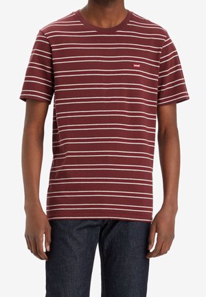 Man draagt een kastanjebruin T-shirt met korte mouwen met dunne witte horizontale strepen en een klein rood Levi’s-logo op de borst, gecombineerd met donkere jeans.