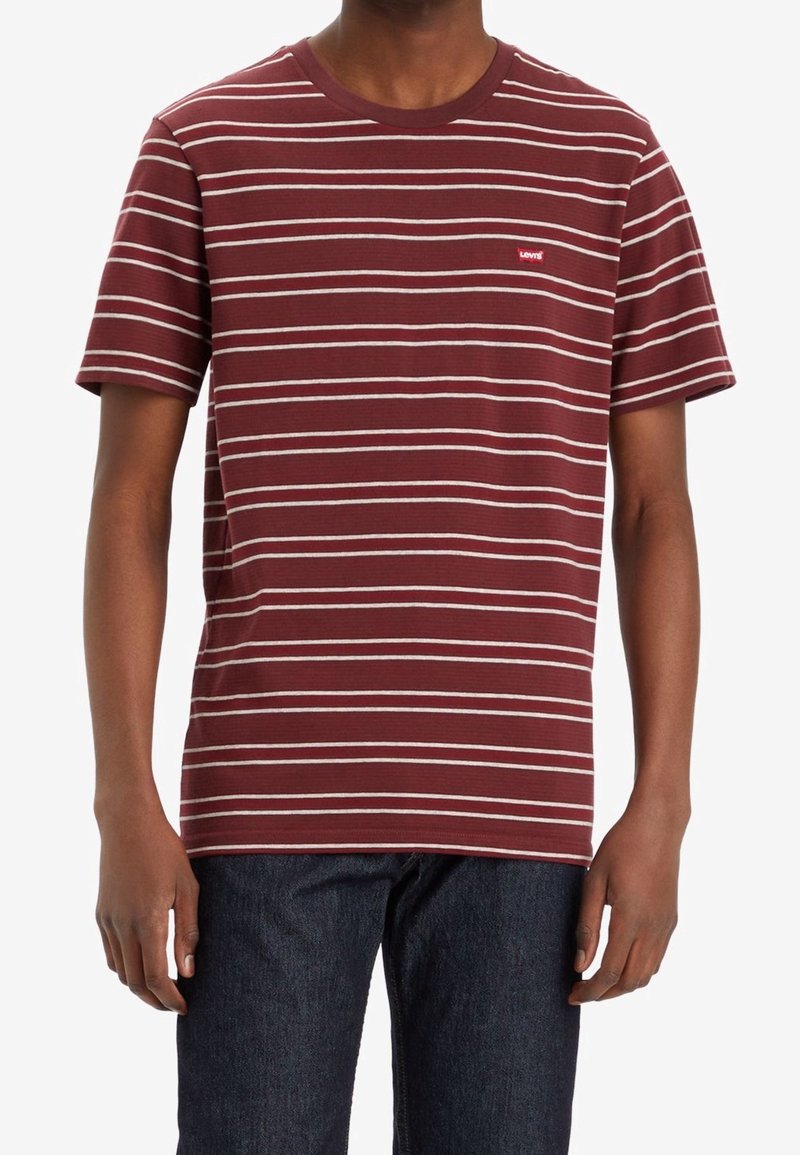 Homme portant un t-shirt à manches courtes bordeaux avec de fines rayures horizontales blanches et un petit logo Levi’s rouge sur la poitrine, associé à un jean foncé.