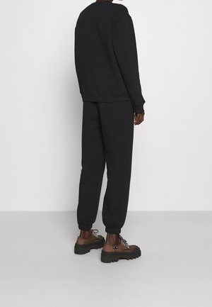 Persoon gekleed in een zwarte top met lange mouwen en zwarte joggerbroek, gecombineerd met bruine veterboots met dikke zolen, staand tegen een witte achtergrond.