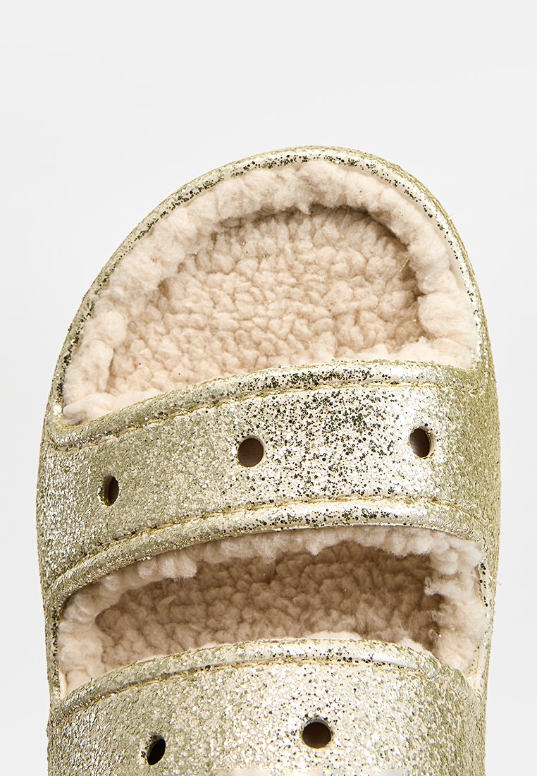 Gouden glinsterende schuifsandalen met twee brede banden, zachte beige bontvoering en open teen ontwerp. Gelaagd oppervlak met ventilatiegaten.