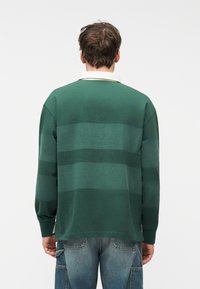 Άνδρας με την πλάτη στραμμένη, φοράει πράσινο ριγέ μακρυμάνικο πουκάμισο με λευκό γιακά και μπλε cargo τζιν με τσέπες.