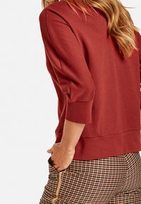 Bordeauxfarbene, leichte Sweatshirt mit 3/4-Ärmeln und abgerundetem Saum, kombiniert mit gemusterten Karohosen mit einem seitlichen Reißverschluss.