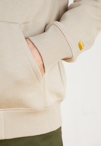 Beige hoodie med ribbade muddar och nederkant, med en stor framficka och en liten broderad gul logotyp på ärmen. Mjuk textur.