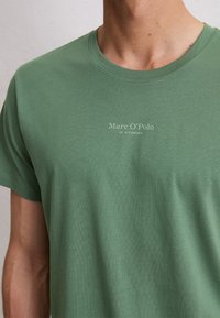 Close-up of person wearing green Marc O’Polo T-shirt with brand name and "EST. IN STOCKHOLM" text on chest.