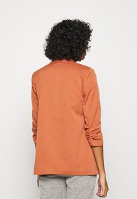 Marynarka w kolorze rdzy o dopasowanym fasonie, z długimi rękawami ze zdobieniem i prostym krojem, wykonana z gładkiej tkaniny.