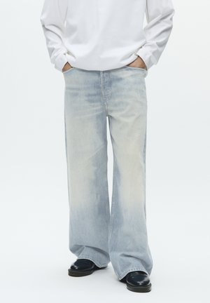 Személy világoskék, bő szárú farmernadrágot, fekete cipőt és fehér, hosszú ujjú inget visel, kezét a zsebében tartja, és fehér háttér előtt áll.