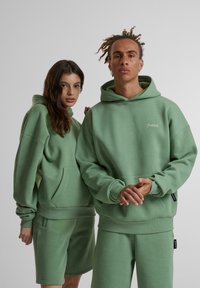 Ensemble de sweat à capuche vert et de shorts avec une coupe décontractée, des poignets côtelés, de grandes poches avant et un détail de logo brodé. Texture douce.