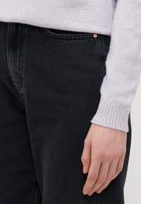 Črne denim jeans s teksturiranim zaključkom, ki vključujejo bakren gumb in vidno šivanje. Deloma je vidna tudi svetlo siva rokav.