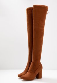 Bottes hautes jusqu'aux genoux en daim marron, avec des talons carrés épais et des bouts arrondis, présentées debout sur une surface blanche.