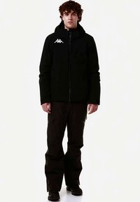 Giacca nera con zip e cappuccio con emblema bianco, abbinata a pantaloni scuri. La giacca presenta una texture liscia e un design aderente.