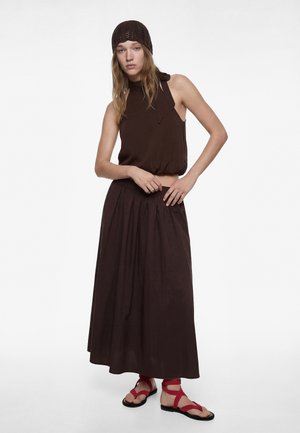 Jonge vrouw met een mouwloze donkerbruine top, bijpassende lange rok, gebreide bruine muts en rode platte sandalen met bandjes, staand tegen een witte achtergrond.