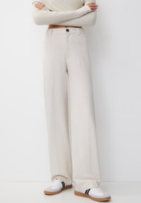 Persoon in hoge taille beige wijde pantalons en witte sneakers met zwarte strepen, staand tegen een witte achtergrond.