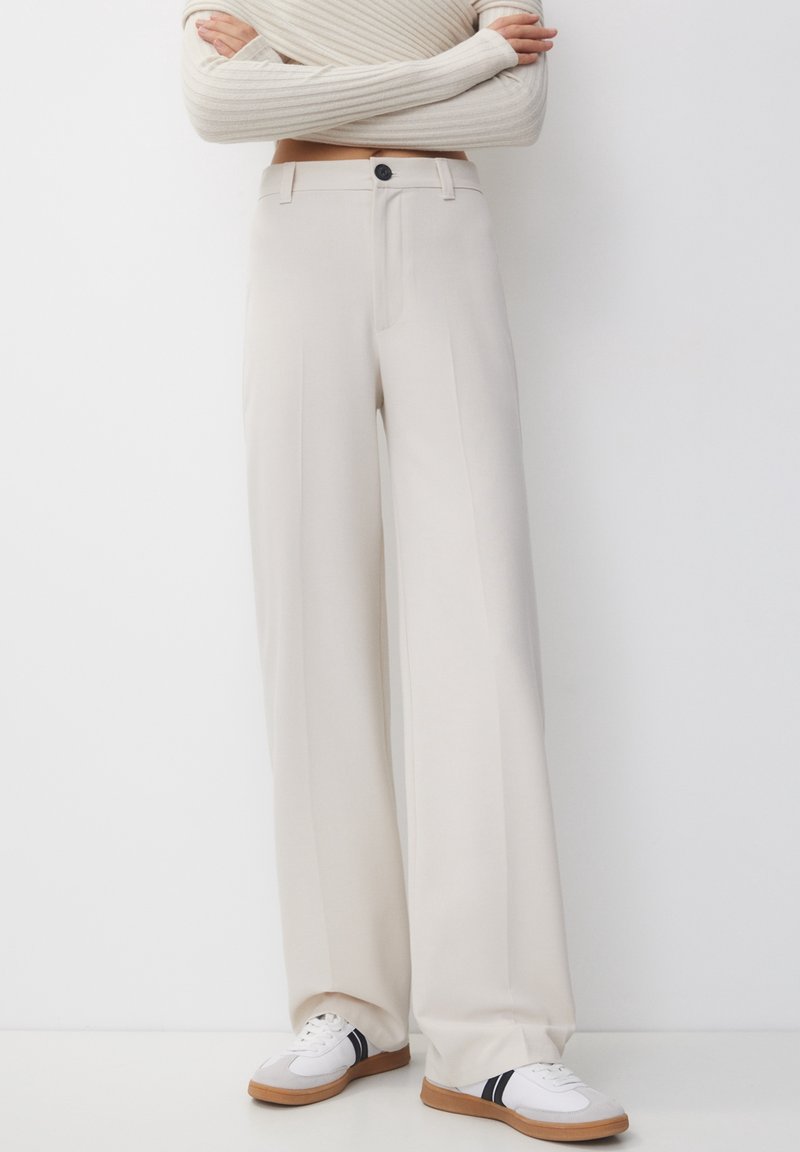 Persoon in hoge taille beige wijde pantalons en witte sneakers met zwarte strepen, staand tegen een witte achtergrond.
