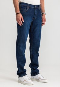 Ciemnoniebieskie dżinsy z prostymi nogawkami, pięcioma kieszeniami i pomarańczowym przeszyciem, noszone z białymi trampkami.