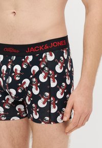 Μαύρα ανδρικά μποξεράκια με κόκκινη ζώνη μέσης, διακοσμημένα με χριστουγεννιάτικο μοτίβο χιονάνθρωπου σε λευκό, κόκκινο και μαύρο. Φτιαγμένα από ελαστικό ύφασμα για άνεση.