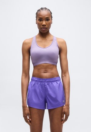 Sportliche Frau mit geflochtenem Haar, die einen fliederfarbenen Sport-BH und lila Laufshorts trägt und vor einem schlichten Hintergrund steht.