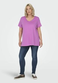T-shirt a manica corta con scollo a V in misto cotone rosa chiaro, abbinato a jeans skinny blu e sneaker marroni con strisce nere.