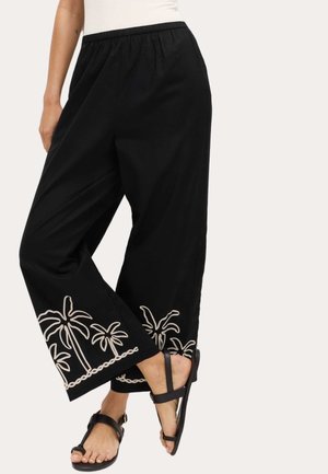 Schwarze Hosen mit weitem Bein und weißer Palmenstickerei an den Bündchen, kombiniert mit schwarzen Riemchensandalen.