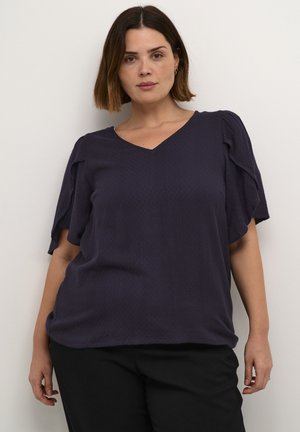 Top de manga corta y escote en V en azul marino profundo con un patrón de diamantes texturizado; corte holgado y mangas ligeramente abullonadas, combinado con pantalones oscuros.