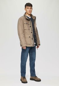 Tarnfarbene, mehrfarbige Jacke mit hohem Kragen über einem dunklen Pullover, kombiniert mit blauen Jeans und braunen Schnürstiefeln.
