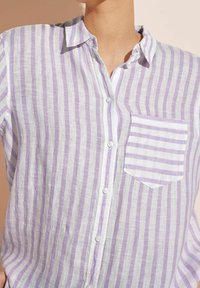 Camicia leggera in lino a righe verticali lilla e bianche. Presenta un colletto, chiusura frontale con bottoni e una tasca sul petto con dettagli a righe.