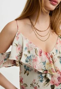 Blusa floreale con maniche scoperte, caratterizzata da uno sfondo chiaro con motivi floreali rosa e blu. Accentata da collane d'oro a strati.