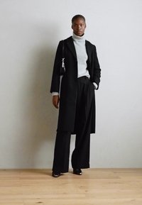 Črn dolg plašč prek sivega turtleneck puloverja, v kombinaciji s širokimi črnimi hlačami. Drži majhno črno torbico; prevladujejo gladke teksture v obleki.