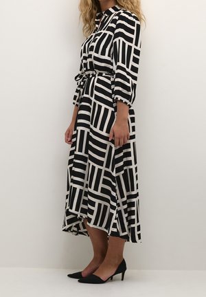 Zwart-wit gestreepte jurk met lange mouwen, een getailleerde taille en een high-low zoom. Draagt zwarte pumps met een spitse neus.