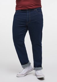 Jeans din denim de culoare albastru închis, cu croială dreaptă, având mansete răsucite și cusături contrastante de culoare portocalie, asortați cu adidași albi.