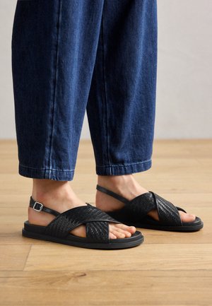 Voeten met zwarte gevlochten leren sandalen met enkelbandjes, gecombineerd met blauwe spijkerbroek op een houten vloer.