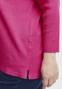 Suéter de punto fucsia con detalle de abertura lateral, mangas largas y ribete acanalado. Se aprecia una textura suave en la superficie de la tela.
