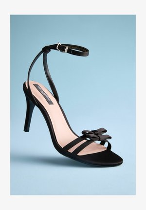 Schwarze hochhackige Sandale mit eleganter Form, Satinschäuften, offener Zehenpartie und einer dekorativen Schleifendetail an der Vorderseite. Die Innensohle ist beige.