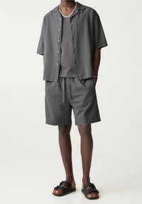 Camisa de botones de manga corta gris sobre una camiseta sin mangas gris, combinada con pantalones cortos gris a juego y sandalias negras con detalles de hebilla.