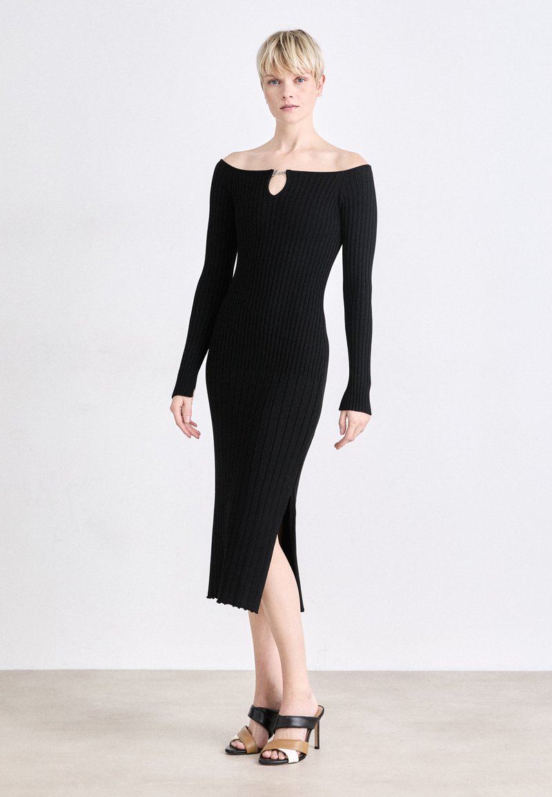 Ribbed black dress with off-shoulder design, keyhole detail at neckline, long sleeves, and side slit. Worn with black and tan high-heeled sandals.