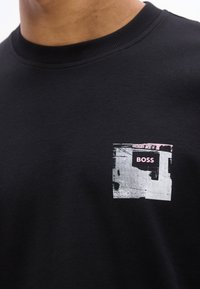 Μαύρο πουκάμισο με στρογγυλή λαιμόκοψη, με ένα μικρό αφηρημένο σχέδιο και τη λέξη «BOSS» σε ροζ χρώμα στην αριστερή πλευρά του στήθους.