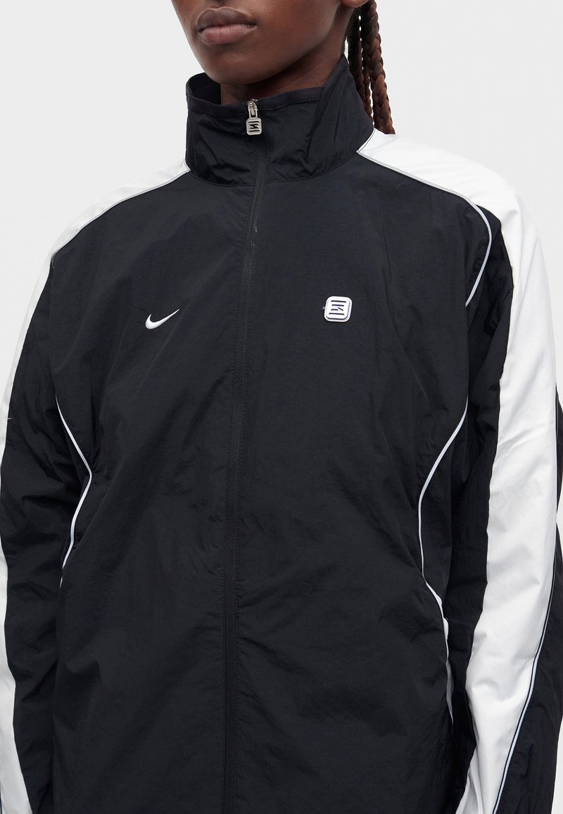 Persoon draagt een zwart-wit Nike trainingsjack met rits tot aan de hoge kraag, met een klein logo op de borst.