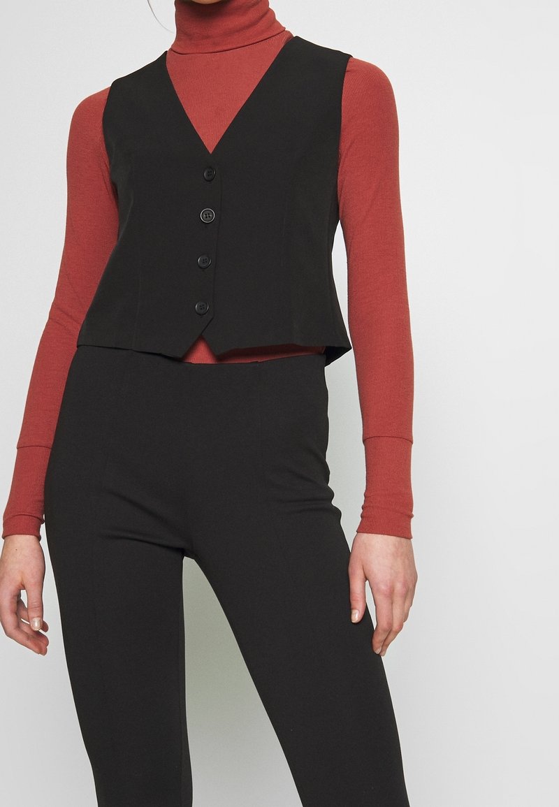 Femme portant un gilet noir boutonné sur un col roulé à manches longues de couleur rouille et un pantalon noir ajusté, debout devant un fond uni.