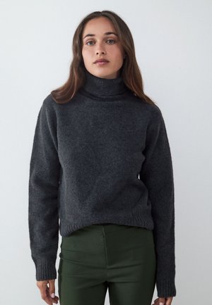 Mujer con cabello largo y castaño, vestida con un suéter de cuello alto gris oscuro y pantalones verde oliva, de pie contra un fondo claro y liso.