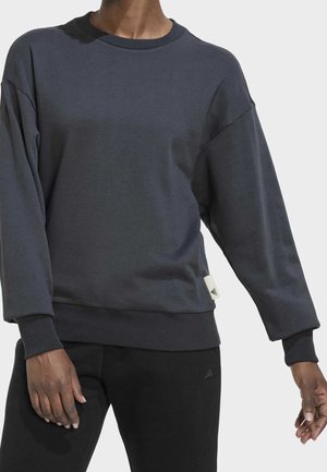Person iført en mørkegrå langærmet sweatshirt og sorte bukser mod en ensfarvet lys baggrund.
