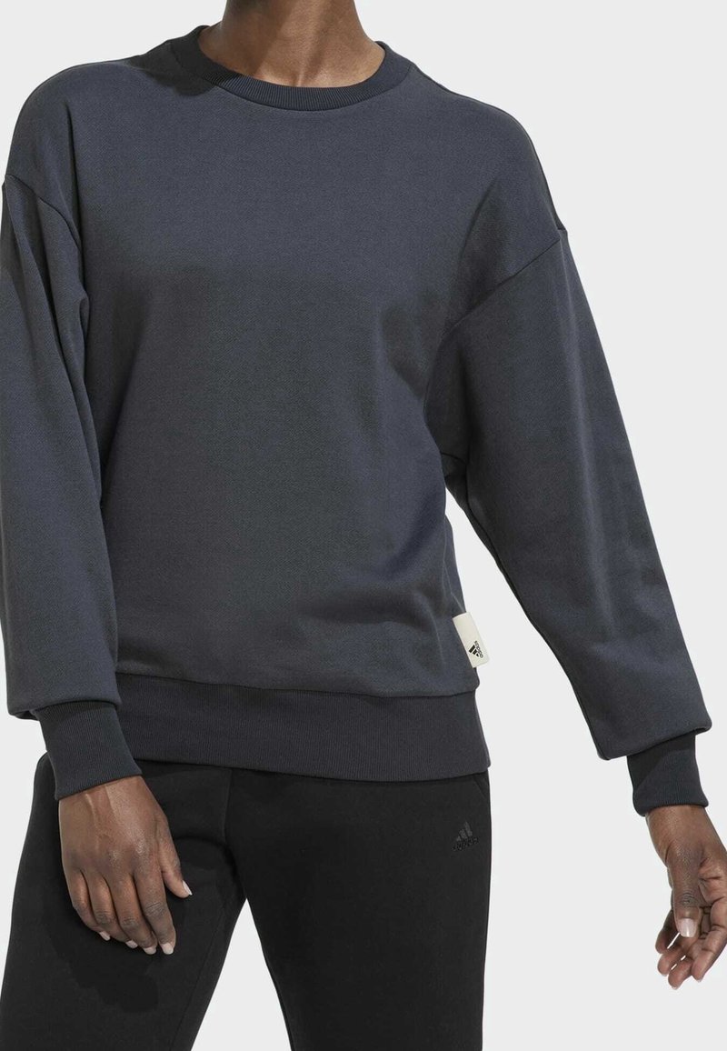 Personne portant un sweat-shirt gris foncé à manches longues et un pantalon noir sur un fond clair uni.