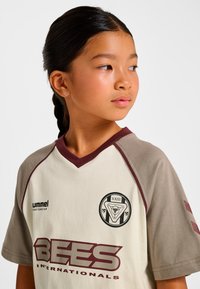 Cremefarbenes Kurzarmtrikot mit kastanienbraunen Akzenten, V-Ausschnitt und einem Logo mit Dreiecksdesign. Das Shirt hat eine lockere Passform.