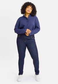 Top de manga larga texturizado en color navy con puños con volantes y detalles de botones, combinado con jeans ajustados oscuros de mezclilla y zapatillas blancas.